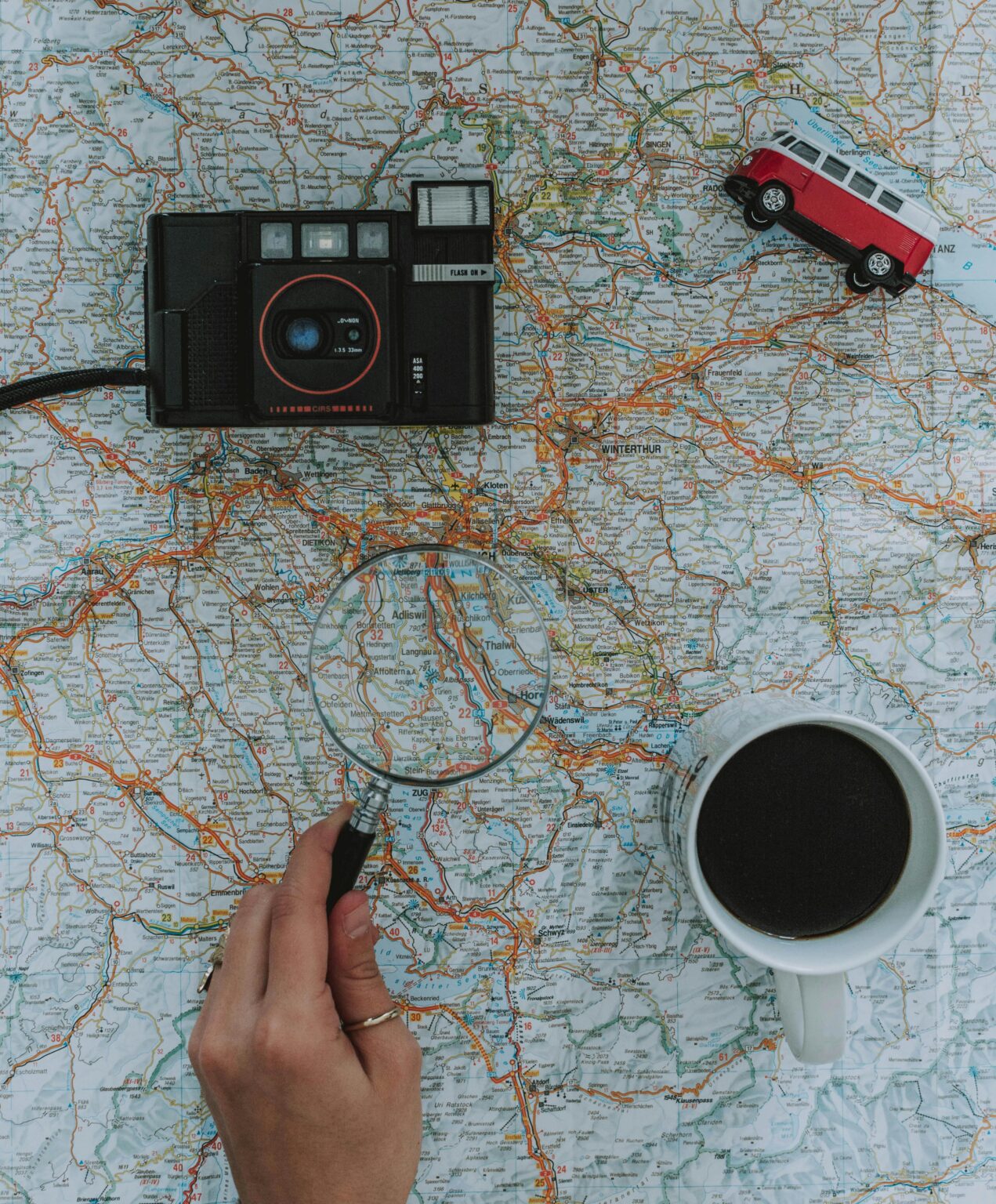 Flat lay of travel essentials: map, camera, toy van, magnifying glass, and coffee.